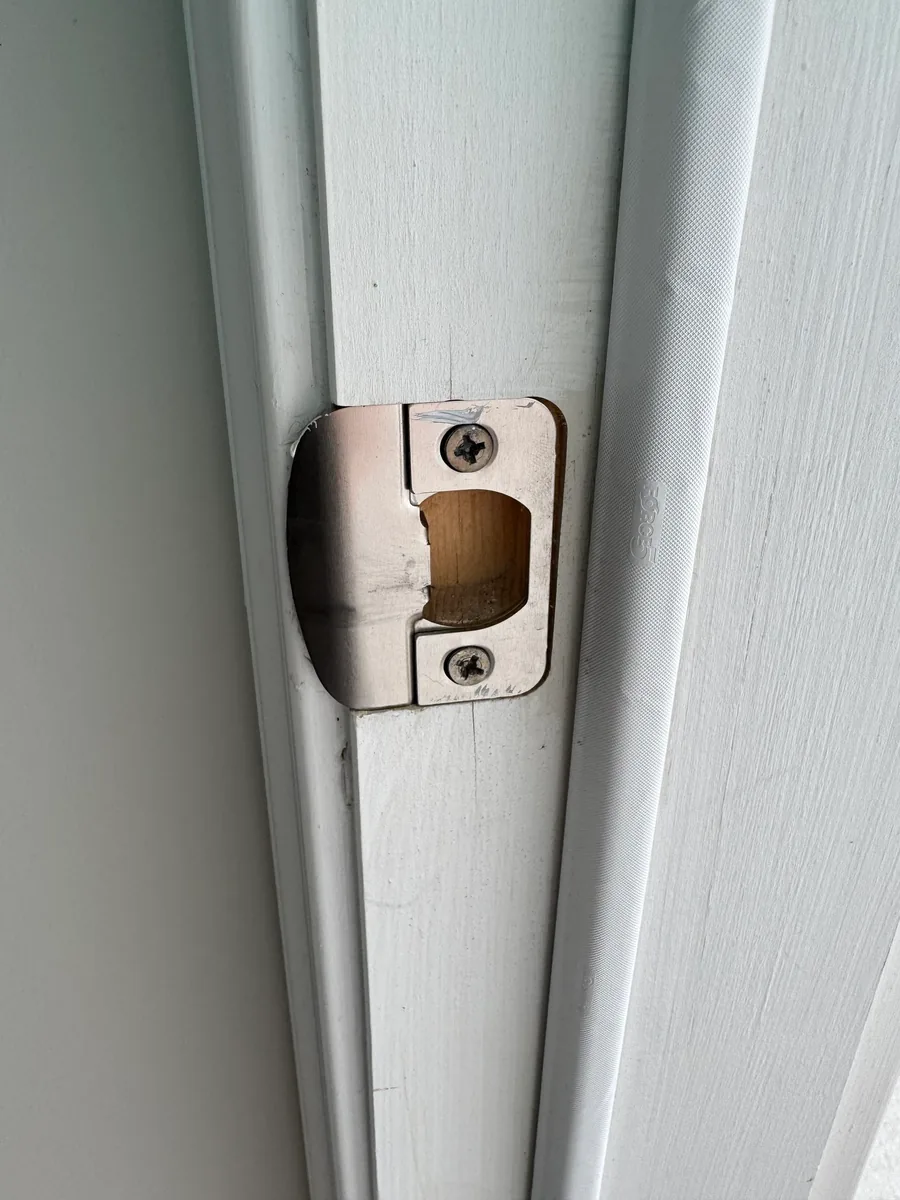 A close-up of a latch strike plate on a door jamb with the mortise behind it opened out to bare wood.